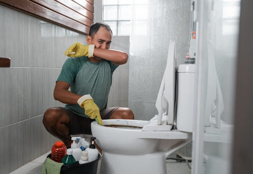 A man covering his nose due to a smelly toilet; he has cleaning supplies with him.